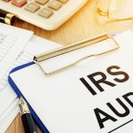 A clipboard with IRS audit paperwork sits beside tax forms, pens, a calculator, and eyeglasses on a desk.