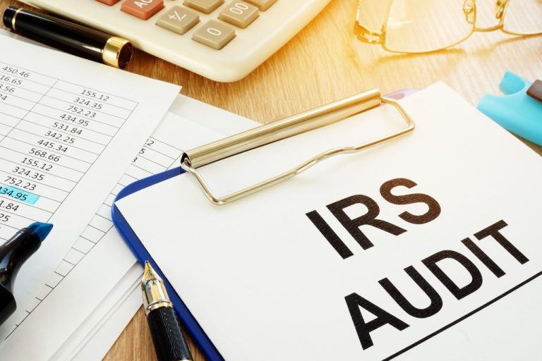 A clipboard with IRS audit paperwork sits beside tax forms, pens, a calculator, and eyeglasses on a desk.