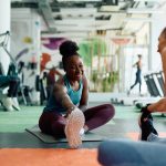 A woman stretching one leg on a mat while another woman sits nearby in a bright gym with exercise equipment.