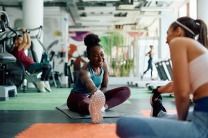 A woman stretching one leg on a mat while another woman sits nearby in a bright gym with exercise equipment.