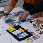 Two people are standing over a desk, working. The desktop is littered with papers, notes, and design mockups.