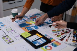 Two people are standing over a desk, working. The desktop is littered with papers, notes, and design mockups.