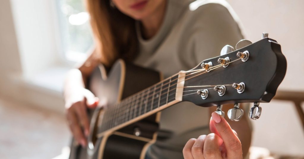 A close-up of a woman sitting in a chair, holding a black guitar, and tuning the guitar strings in a bright room.