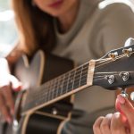 A close-up of a woman sitting in a chair, holding a black guitar, and tuning the guitar strings in a bright room.