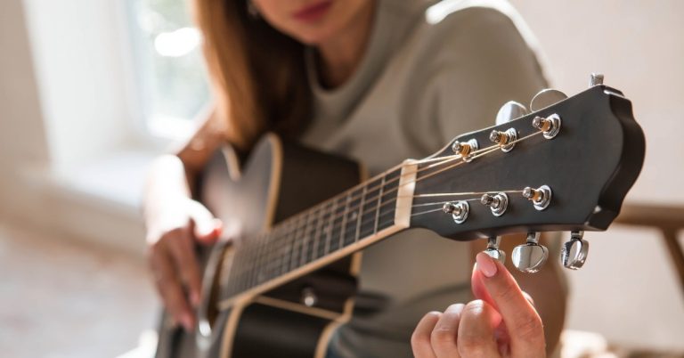 A close-up of a woman sitting in a chair, holding a black guitar, and tuning the guitar strings in a bright room.