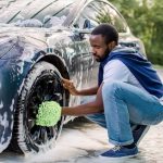 A man crouching beside a soapy car while scrubbing a front wheel with a green microfiber wash mitt.