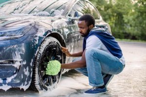 A man crouching beside a soapy car while scrubbing a front wheel with a green microfiber wash mitt.