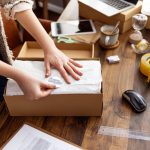 A close-up of a woman packing products into cardboard boxes at a table with other shipping supplies nearby.