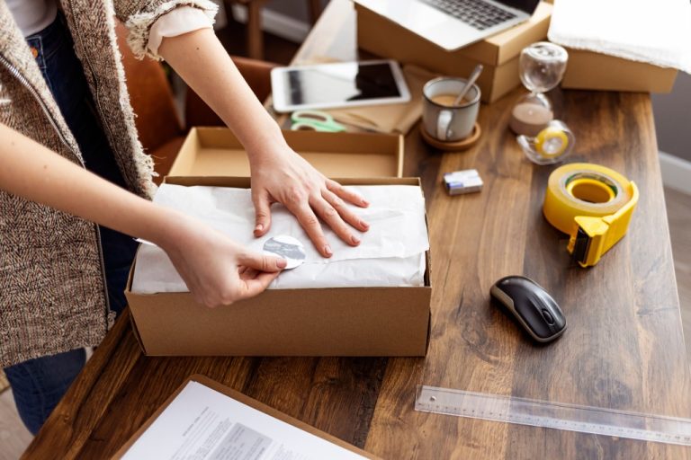 A close-up of a woman packing products into cardboard boxes at a table with other shipping supplies nearby.