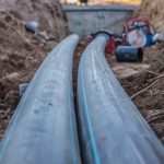 Two gray pipes extending through a dirt trench with loose soil, roots, and construction tools in the background.