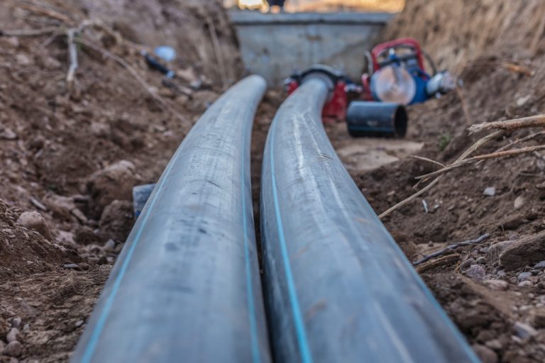 Two gray pipes extending through a dirt trench with loose soil, roots, and construction tools in the background.